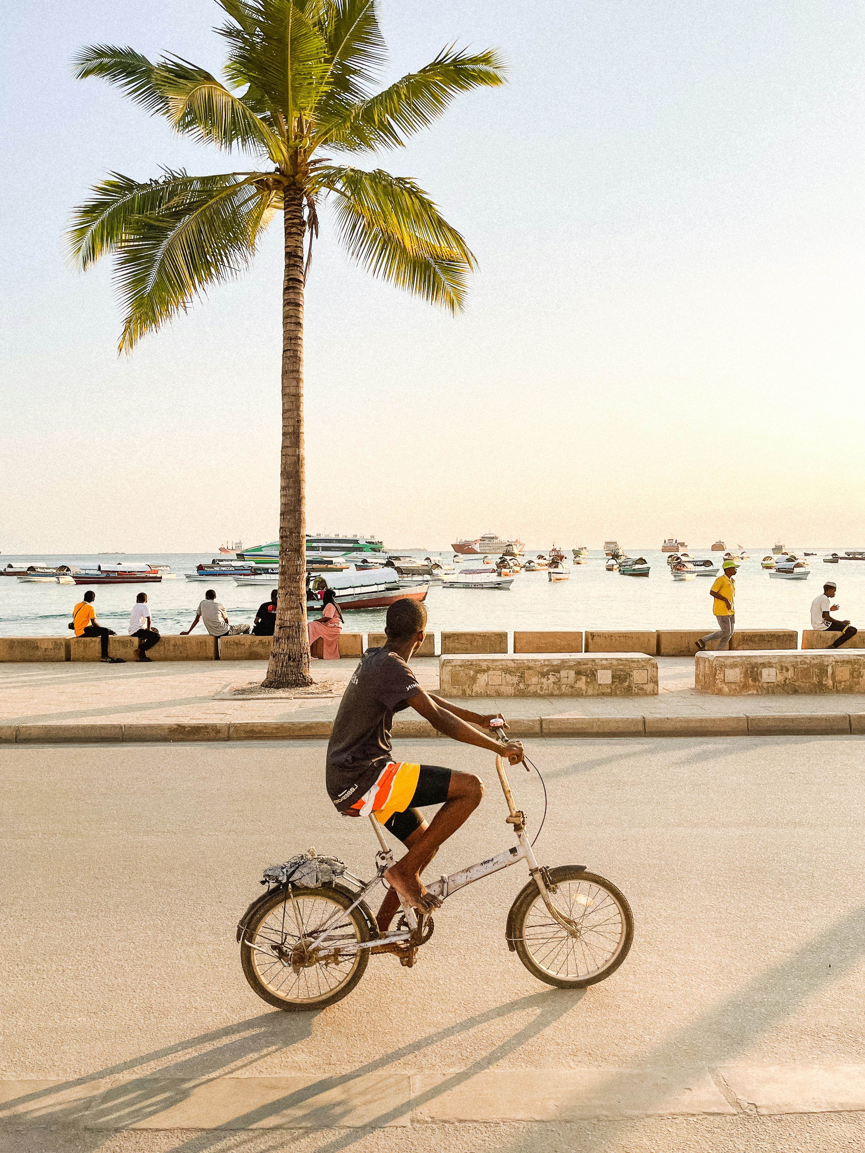 Young Man Riding a Bicycle Along the Coast Looking at the Boats Anchored by the Shore