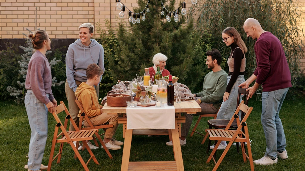 Happy family members talking and sitting down to eat tasty food at big wooden table in backyard in daytime